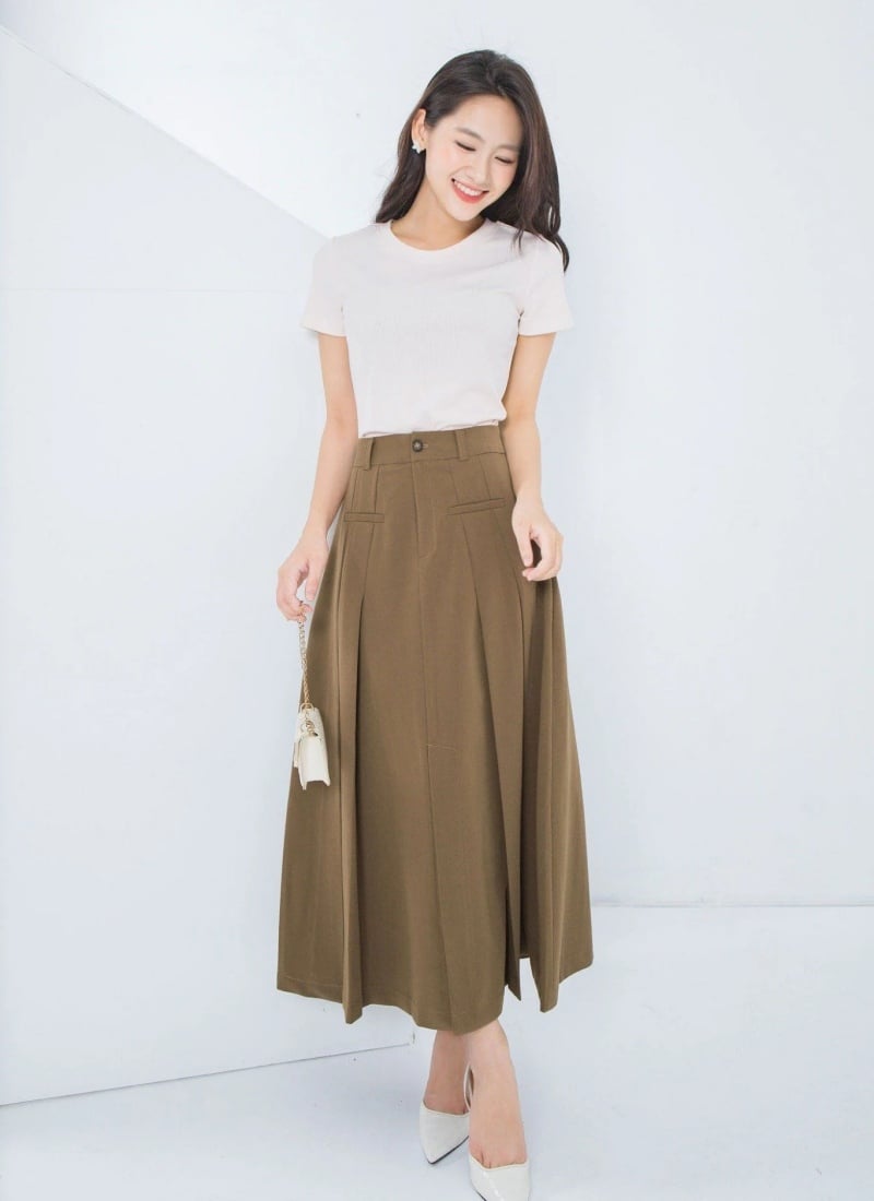 Người phụ nữ mặc áo trắng và váy dài màu nâu, cầm túi xách nhỏ, đứng trong không gian sáng màu.