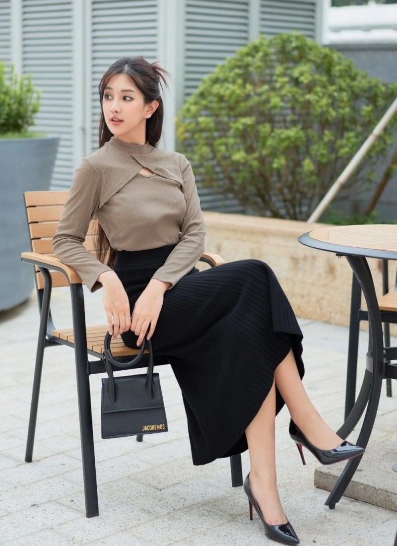 Người phụ nữ ngồi trên ghế ngoài trời, mặc áo dài tay nâu, váy đen, cầm túi xách nhỏ và đi giày cao gót.