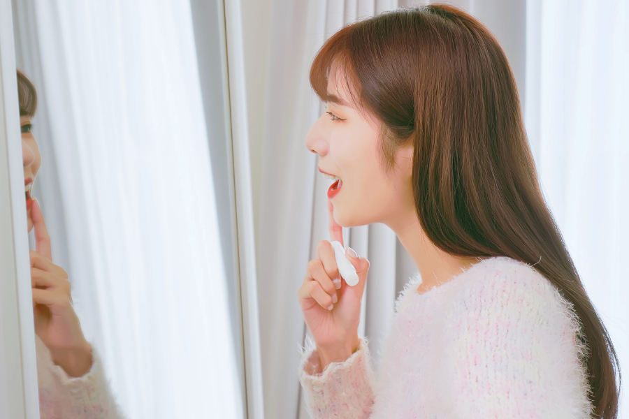 Người phụ nữ soi gương, tay cầm son dưỡng môi, chuẩn bị thoa lên môi trong căn phòng có rèm cửa trắng.