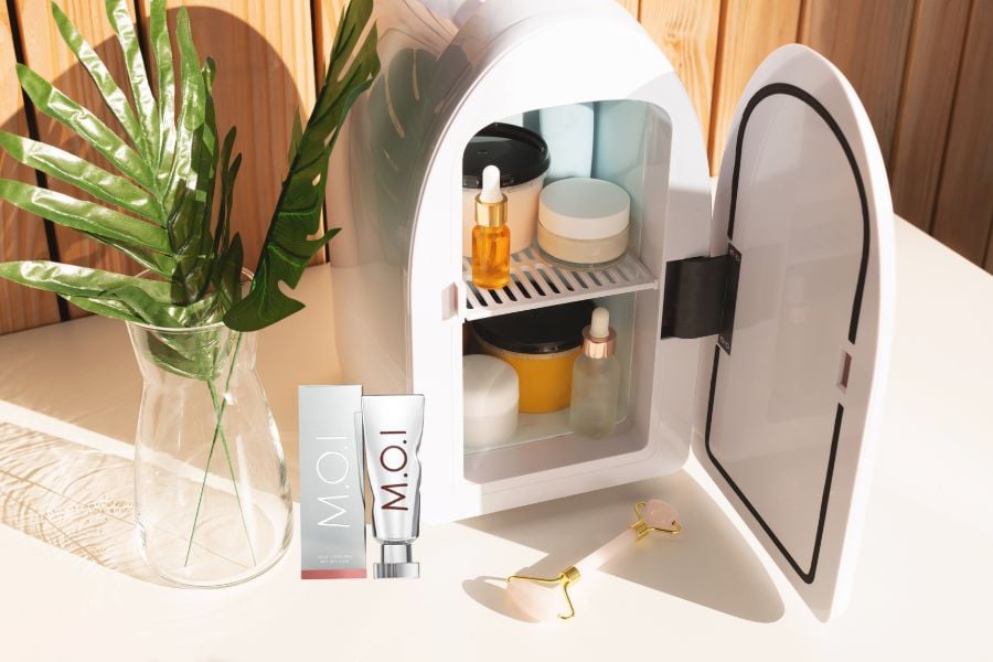 Tủ lạnh nhỏ chứa mỹ phẩm và lọ serum, cạnh đó là cây xanh trong bình và các sản phẩm chăm sóc da.