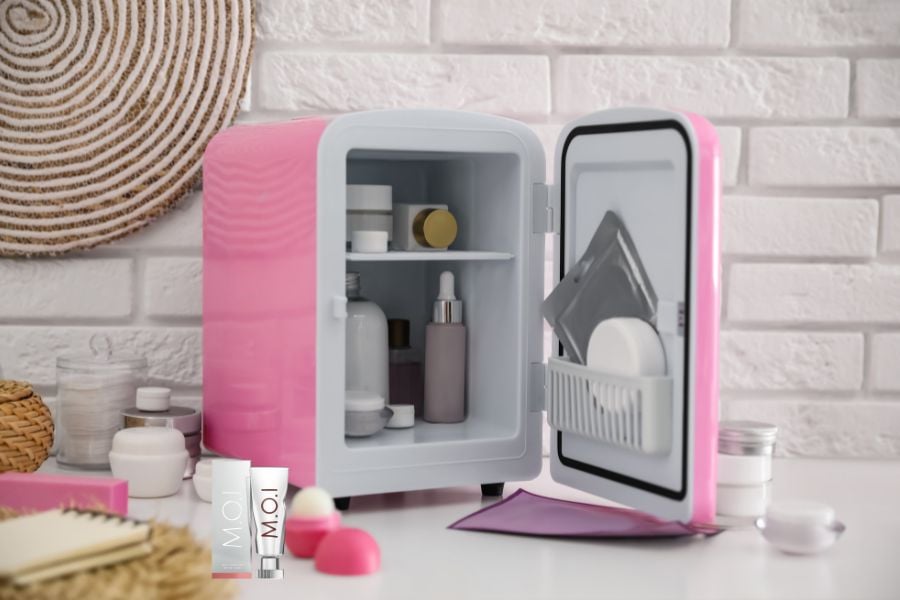 Tủ lạnh mini màu hồng chứa nhiều sản phẩm chăm sóc da, đặt trên bàn trước tường gạch trắng.