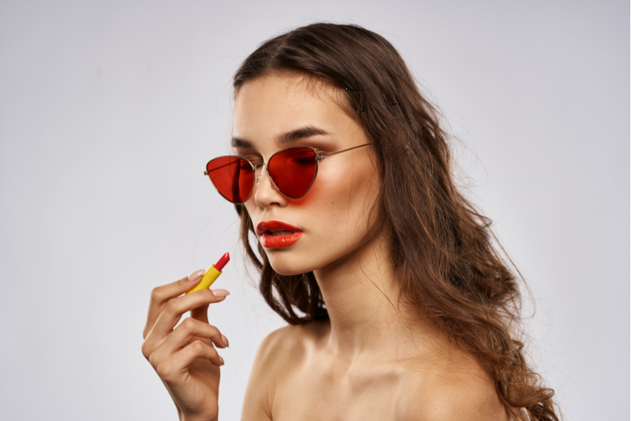 Người đeo kính râm màu đỏ cầm thỏi son đỏ, tóc dài, nền trắng.