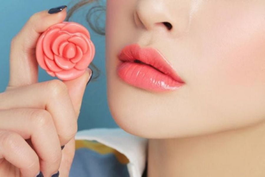 Người cầm son môi hình hoa hồng màu hồng, môi được trang điểm tông màu tương tự.