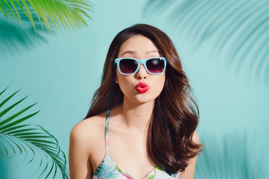 Người đeo kính mát màu xanh, mặc áo họa tiết nhiệt đới, đứng trước nền xanh với cây cọ trang trí.
