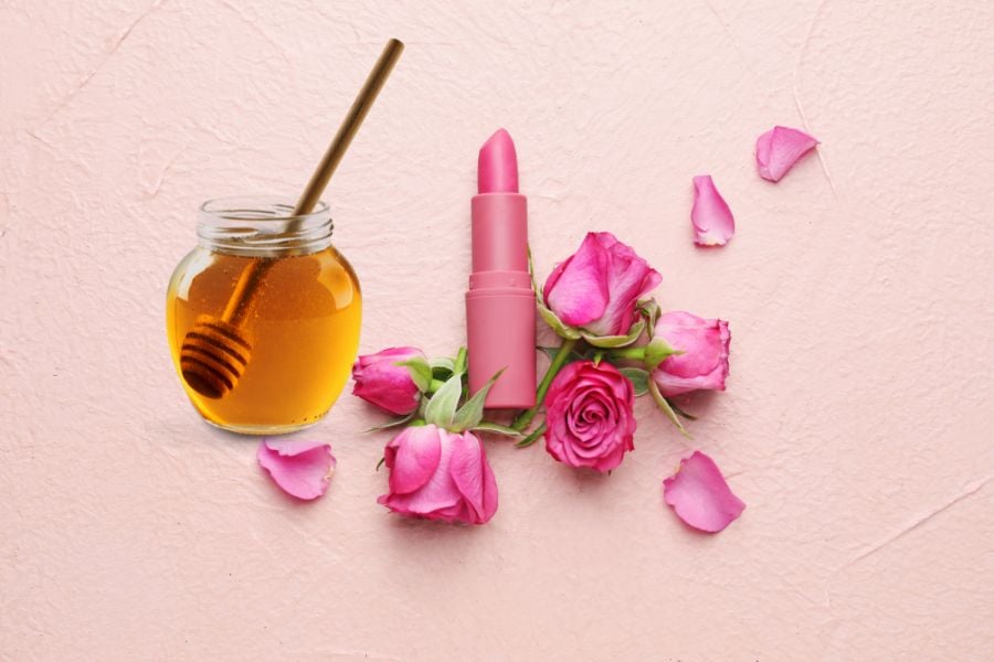 Hũ mật ong và son môi hồng trên nền hồng, xung quanh có hoa hồng và cánh hoa rải rác.