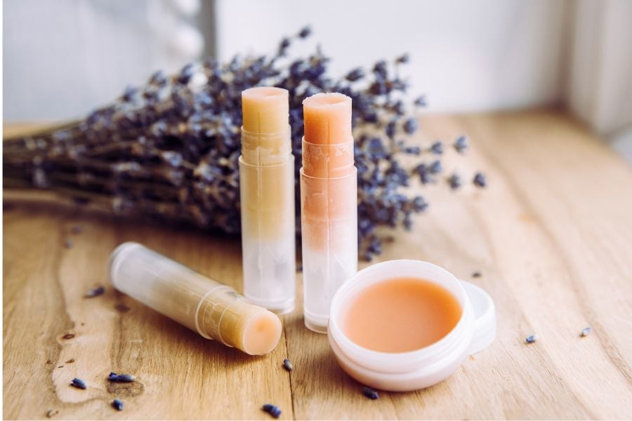 Ba thỏi son dưỡng và một hộp son mở nắp đặt trên bàn gỗ, nền là một bó hoa khô.