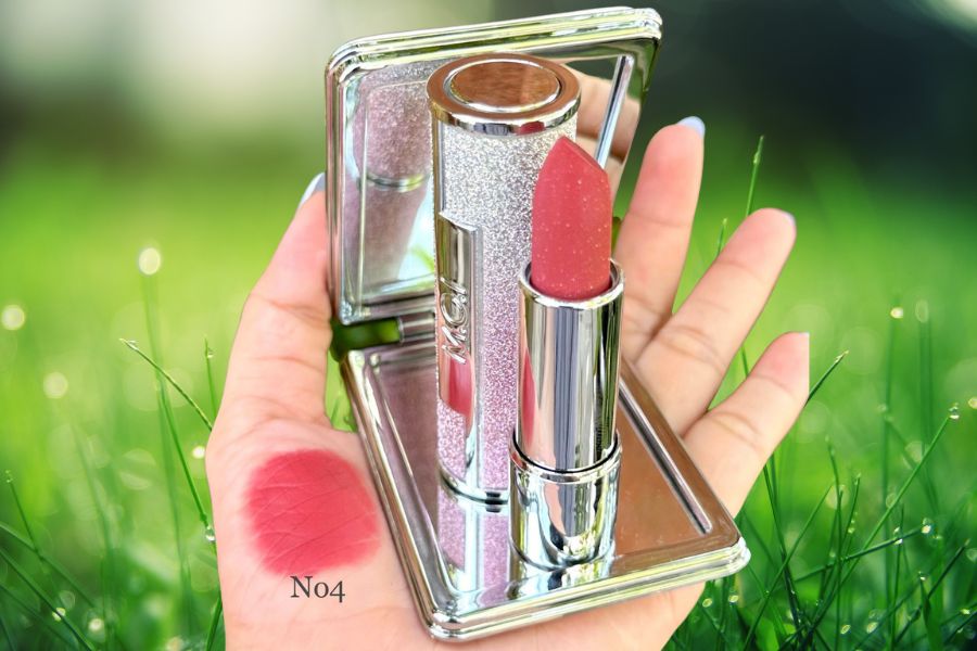 Thỏi son màu hồng cùng vỏ lấp lánh và gương, được cầm trên tay, nền là cỏ xanh mướt.