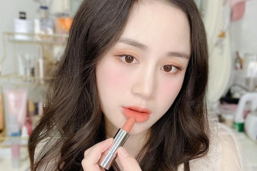Người phụ nữ đang tô son môi màu cam nhạt, tóc dài nâu, đứng trong phòng có nhiều sản phẩm làm đẹp.