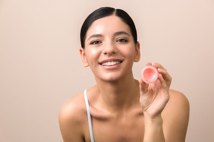 Người phụ nữ mỉm cười, cầm hộp son dưỡng hồng nhỏ, nền màu be.