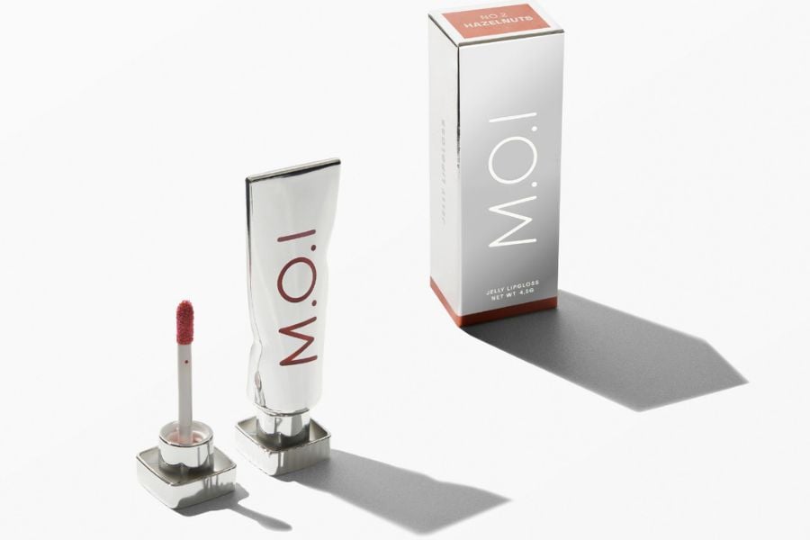 Hộp và tuýp son bóng màu bạc với chữ "M.O.I", có cọ son mở nắp, trên nền trắng.