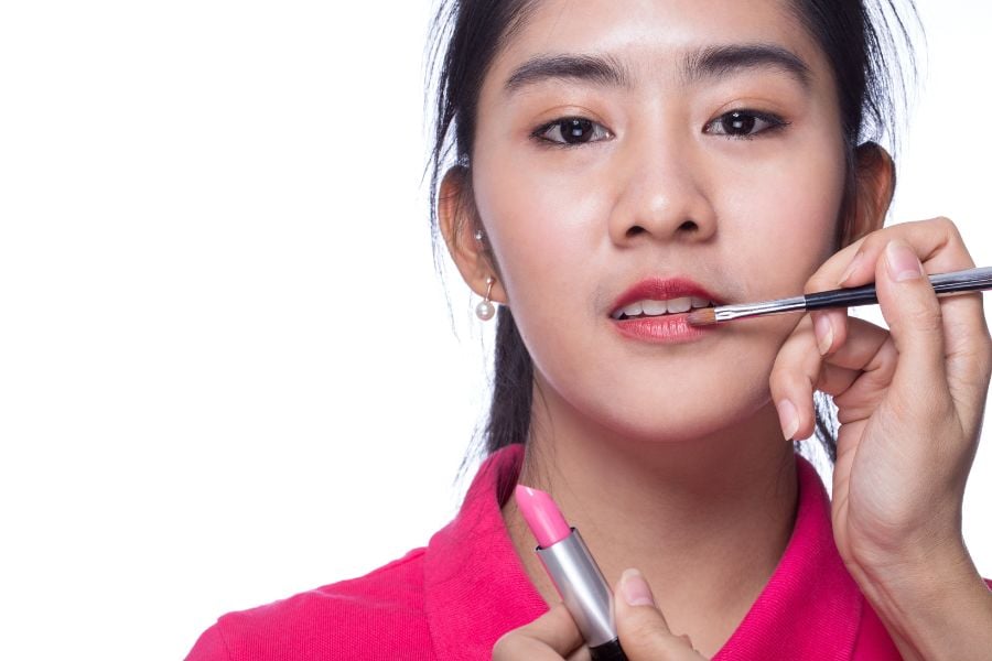 Người đang được trang điểm môi bằng cọ, cầm son môi màu hồng, nền trắng.