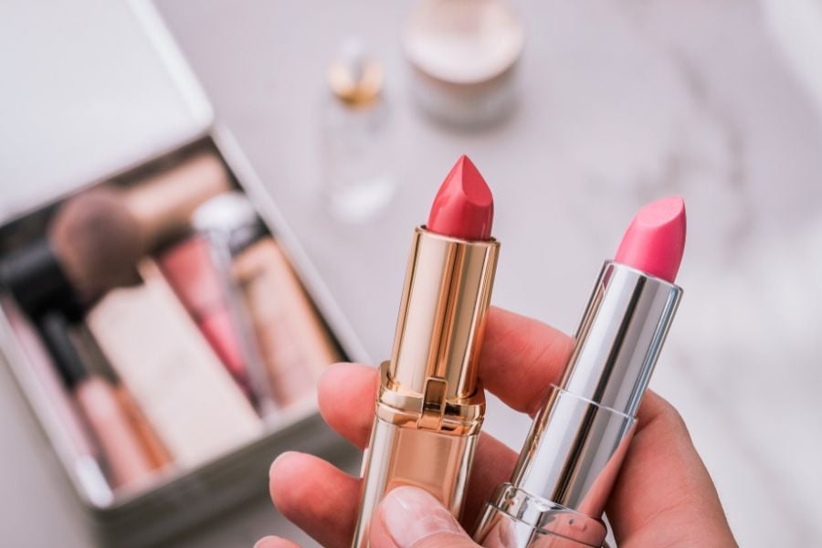 Hai thỏi son màu đỏ và hồng đang được cầm trên tay, nền là hộp đựng đồ trang điểm.
