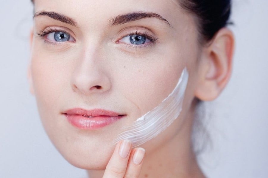 Người phụ nữ thoa kem dưỡng da lên má, gương mặt tươi cười, nền trắng.