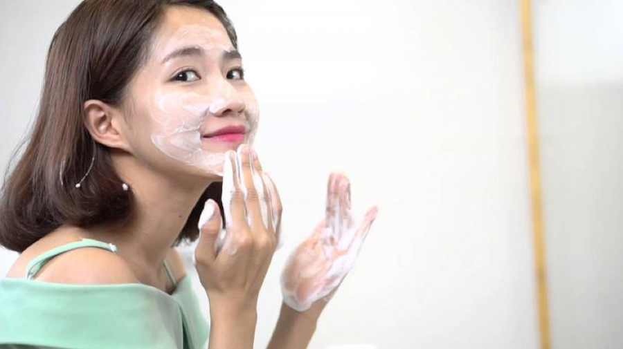 Người phụ nữ đang thoa kem dưỡng da mặt, mặc áo xanh, nền trắng mờ.