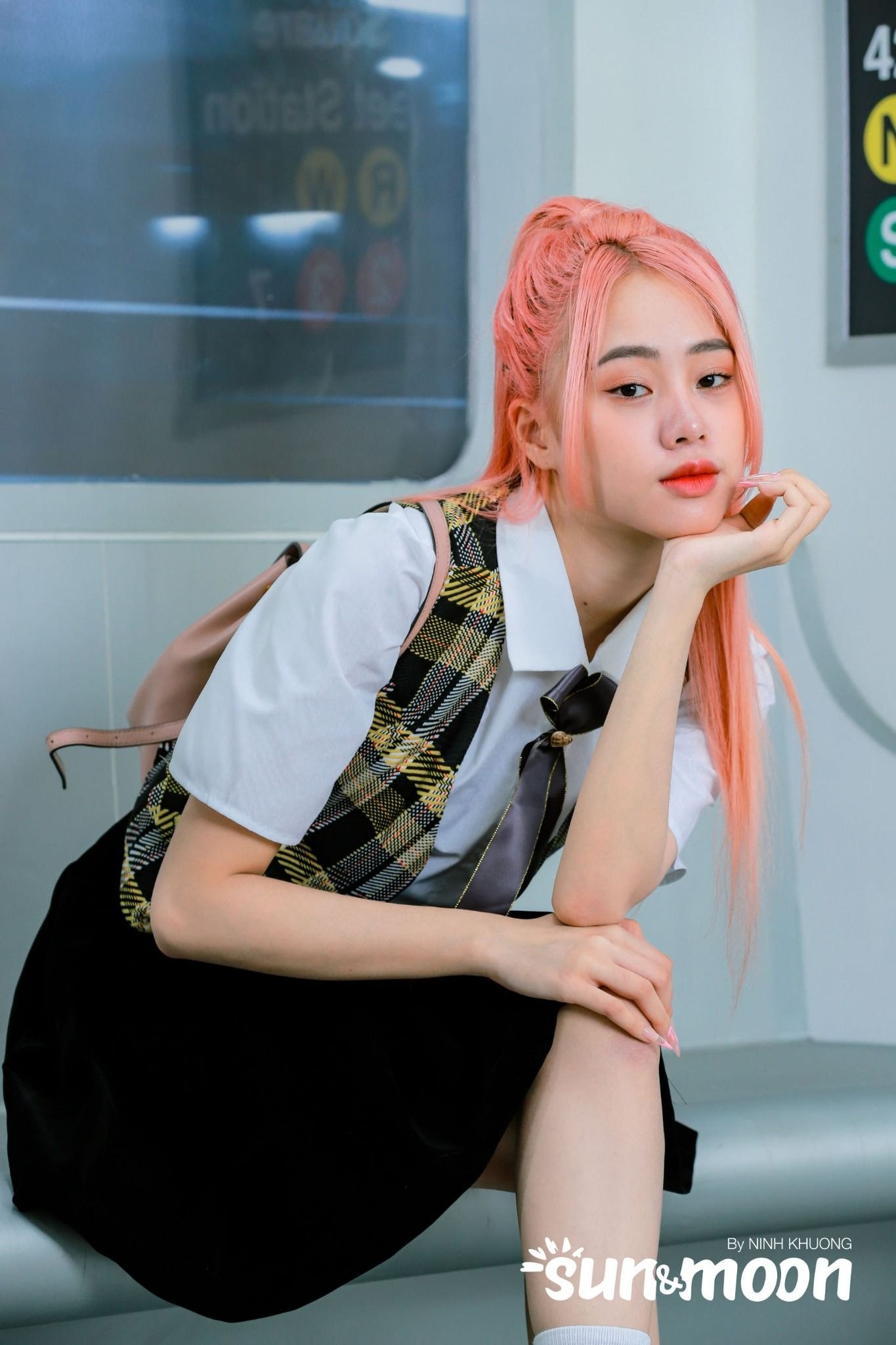 Người có mái tóc hồng, mặc đồng phục học sinh, ngồi trong tàu điện ngầm, tay chống cằm nhìn về phía trước.