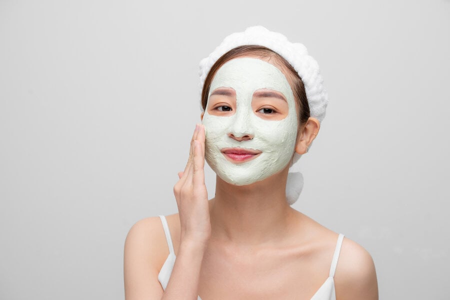Người đang đắp mặt nạ dưỡng da màu xanh, đội khăn trắng, chạm nhẹ vào má, nền trắng.