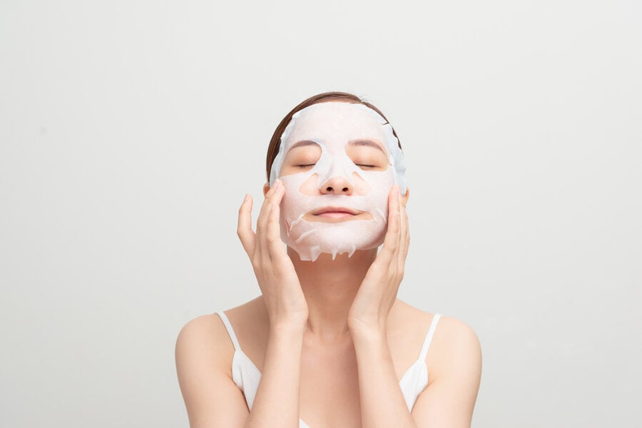 Người đắp mặt nạ dưỡng da, thư giãn với mắt nhắm, tay chạm nhẹ lên mặt, nền trắng.