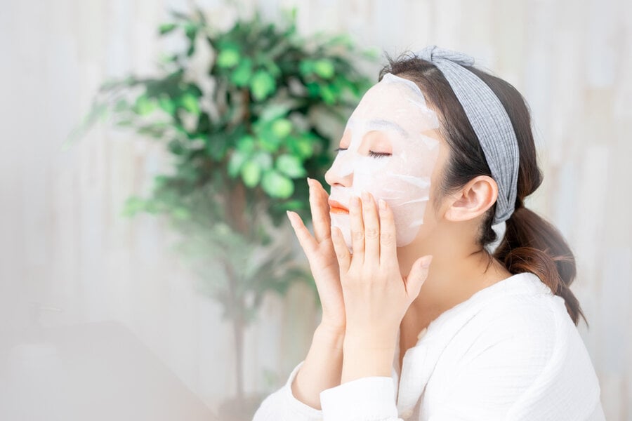 Người đang đắp mặt nạ dưỡng da, mắt nhắm thư giãn, đeo băng đô, nền có cây xanh.