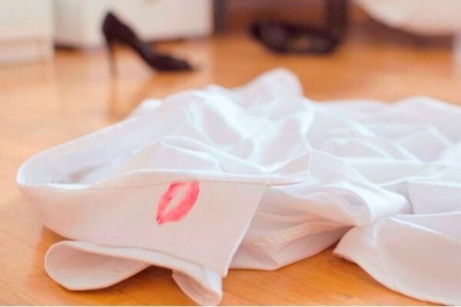 Áo sơ mi trắng trên sàn gỗ với vết son đỏ, giày cao gót đen mờ phía xa.