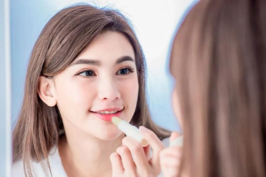 Người phụ nữ soi gương và thoa son dưỡng lên môi, tạo khung cảnh tự nhiên và nhẹ nhàng.