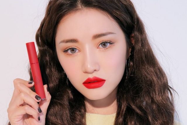 Người phụ nữ cầm thỏi son đỏ, trang điểm với son môi đỏ nổi bật, nền trắng.