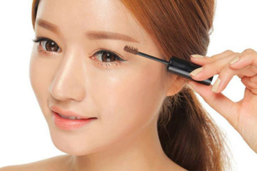 Người phụ nữ đang dùng mascara để chải lông mày, khuôn mặt cận cảnh, tóc nâu buộc gọn phía sau.