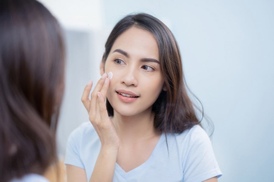 Người phụ nữ thoa kem dưỡng lên mặt, đứng trước gương, mặc áo thun xanh nhạt.