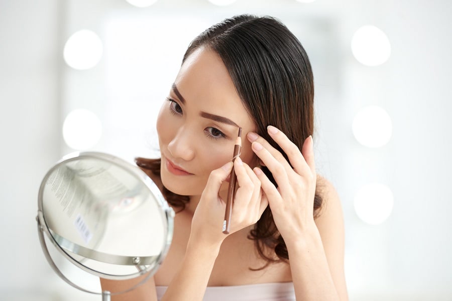 Người phụ nữ đang kẻ lông mày trước gương tròn, trong không gian sáng sủa với ánh đèn nền phía sau.