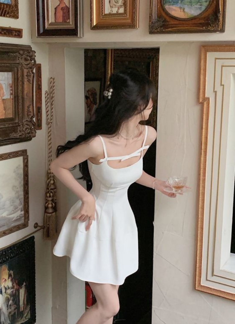 Người mặc váy trắng đứng trước bức tường treo nhiều tranh, tay cầm ly thủy tinh nhỏ.