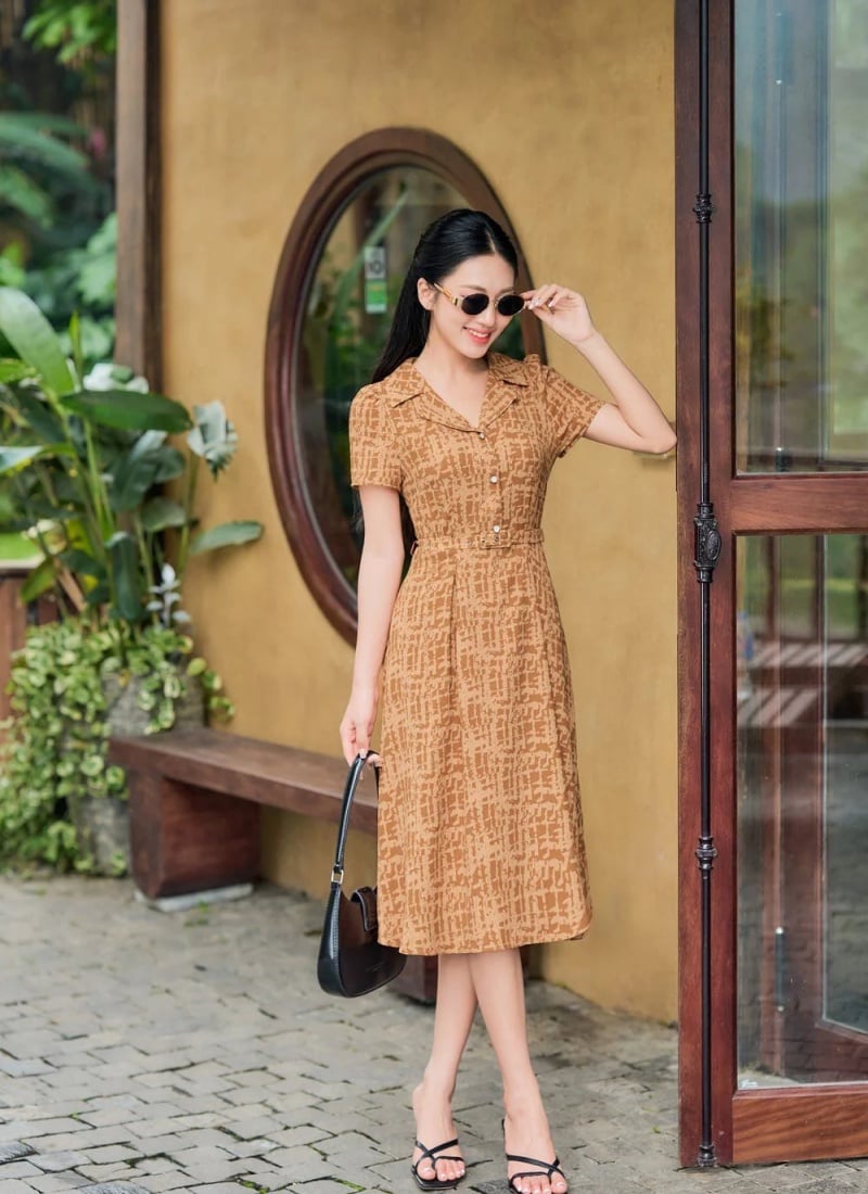 Người phụ nữ mặc váy nâu đứng trước cửa kính, tay cầm kính mát, xung quanh là cây xanh và bức tường vàng.