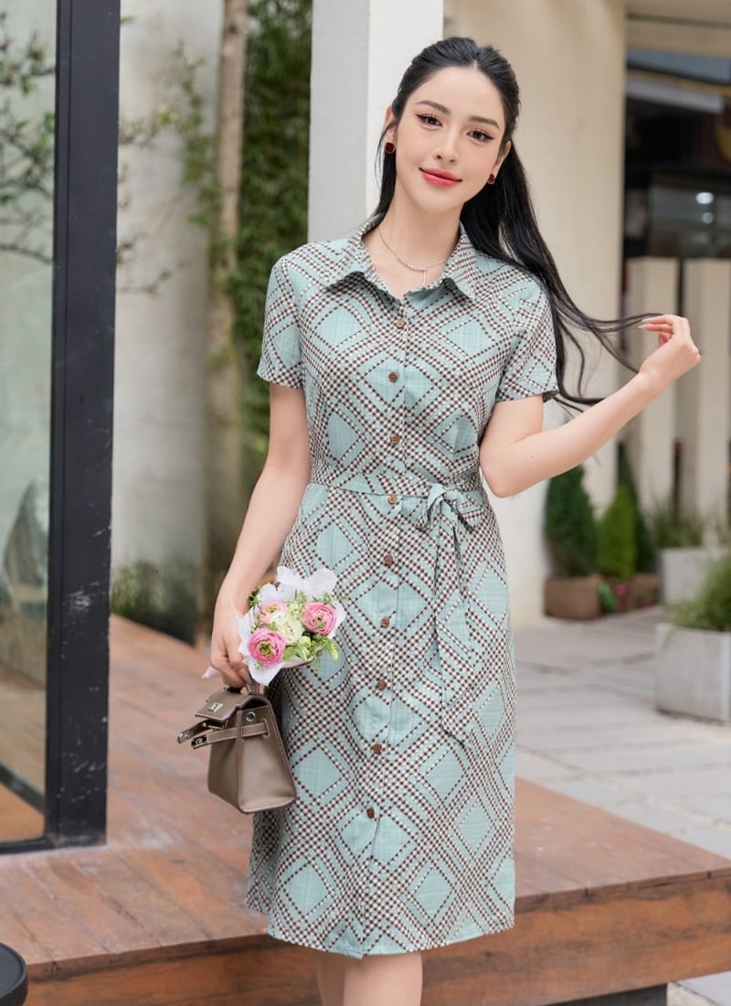 Người phụ nữ mặc váy họa tiết, cầm túi xách và bó hoa nhỏ, đứng trước tòa nhà trắng.
