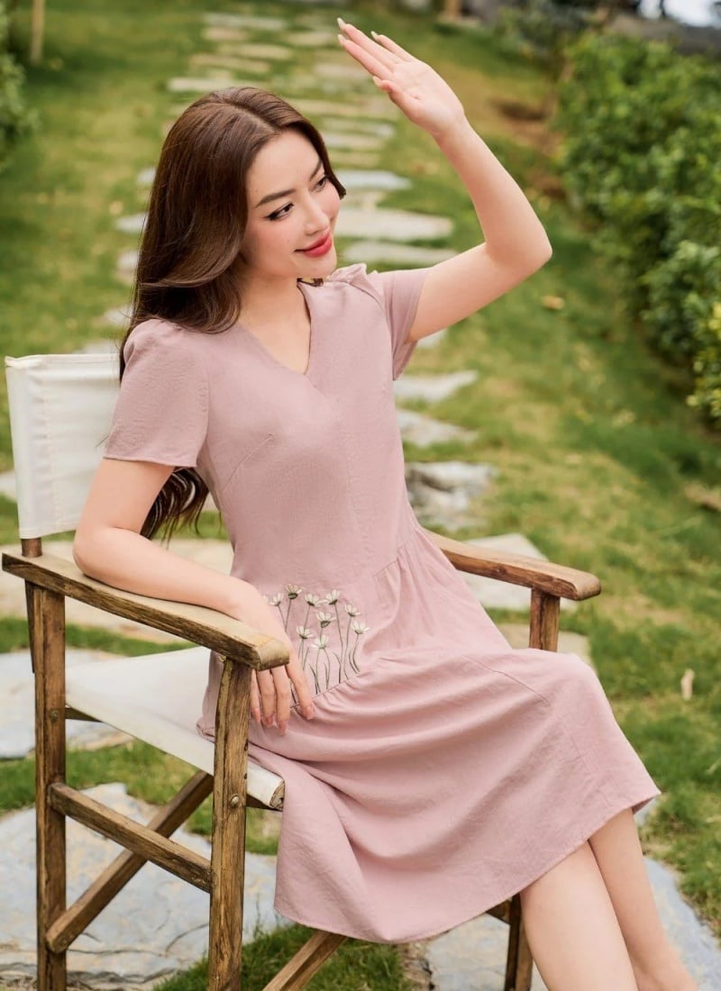 Người phụ nữ trong trang phục hồng ngồi trên ghế ngoài trời, giơ tay che nắng, nền là lối đi và cây xanh.