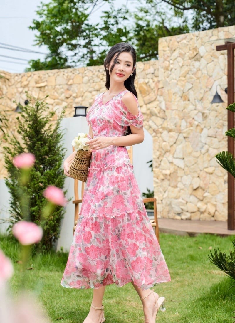 Người phụ nữ mặc váy hoa hồng đứng trong khu vườn, cầm giỏ hoa, nền là bức tường đá và cây xanh.