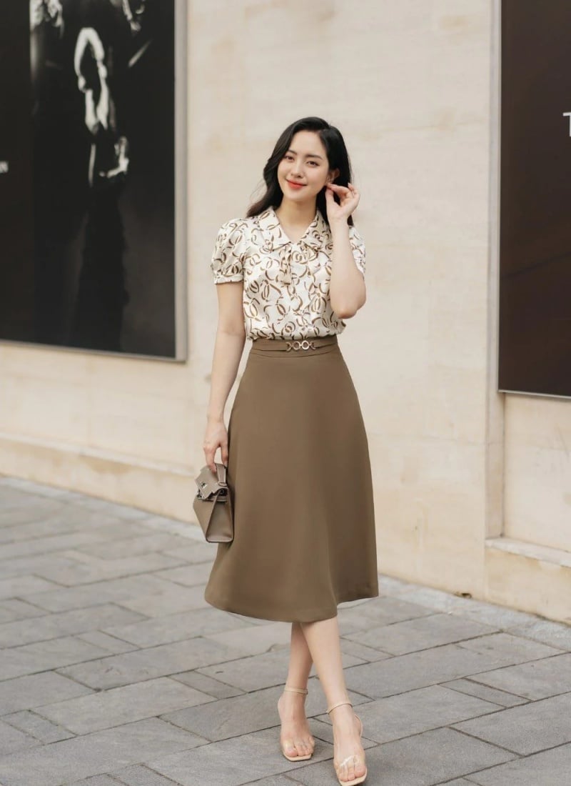 Người phụ nữ mặc áo sơ mi họa tiết và váy nâu đứng trên vỉa hè, cầm túi xách nhỏ, nền tường và bức tranh phía sau.