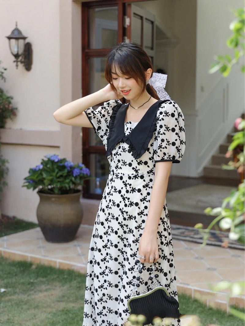 Người phụ nữ mặc váy hoa trắng đen đứng trước cửa nhà, tay cầm túi xách nhỏ, xung quanh có cây cảnh.