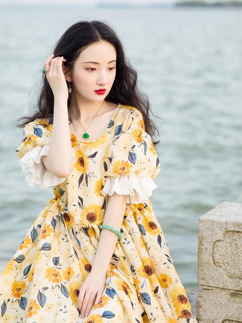 Người mặc váy hoa vàng đứng cạnh bờ nước, tóc dài, tay chạm vào tóc, nhìn xa xăm, phông nền là mặt nước.