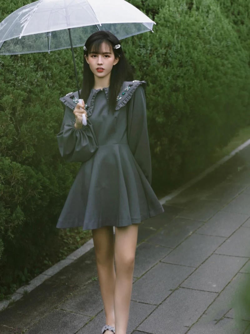 Người cầm ô trong suốt, mặc váy tối màu, đứng trên lối đi lát gạch cạnh hàng cây xanh.