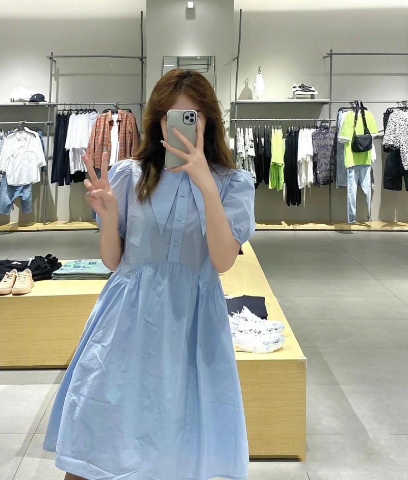 Người mặc váy xanh chụp ảnh trước gương trong cửa hàng quần áo, xung quanh có nhiều trang phục treo trên giá.