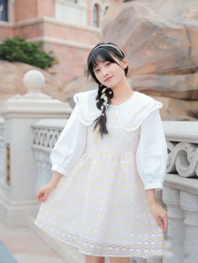 Người phụ nữ mặc váy trắng với họa tiết vàng đứng cạnh lan can, nền là tòa nhà gạch và tảng đá lớn.