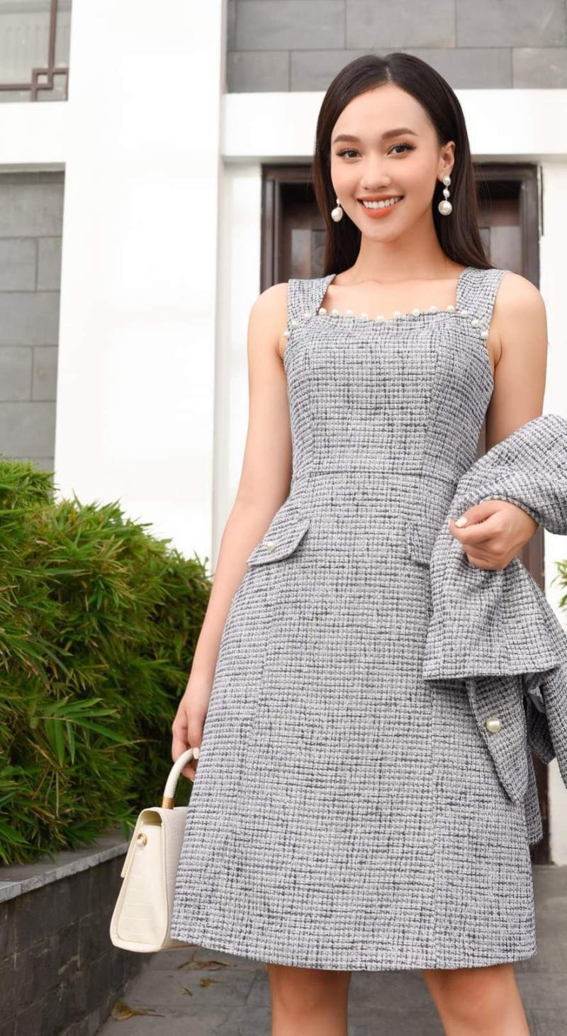 Người phụ nữ mặc váy xám, cầm áo khoác và túi xách trắng, đứng trước tòa nhà với cây xanh xung quanh.