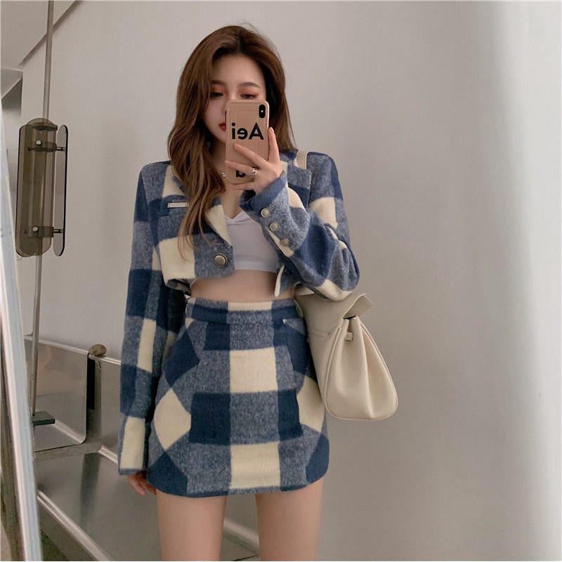 Người đứng trước gương, mặc trang phục caro xanh trắng, cầm điện thoại và mang túi xách màu kem.