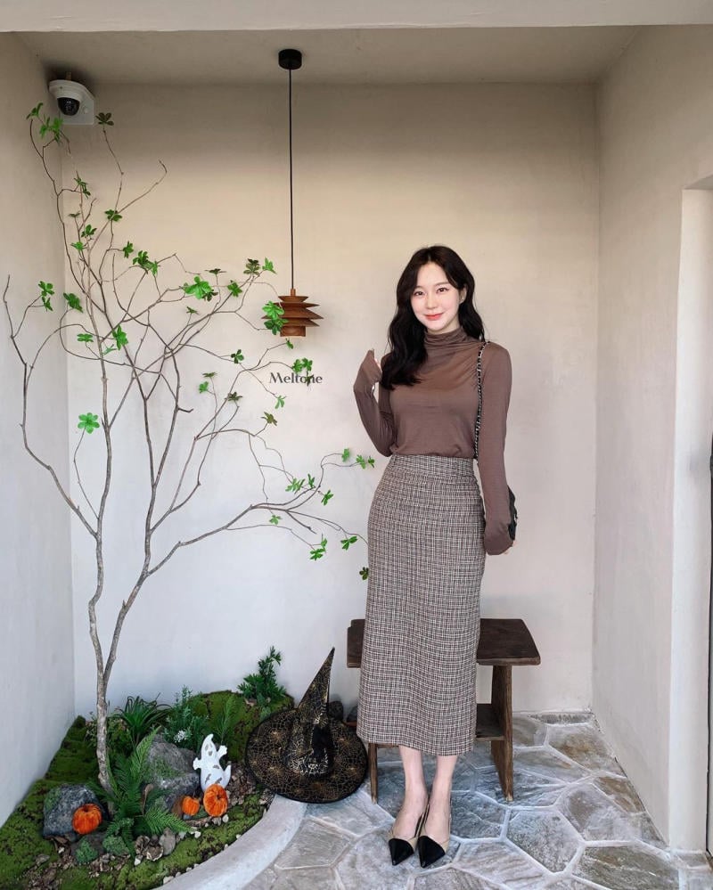 Người đứng cạnh cây trang trí nhỏ trong phòng, mặc áo dài tay nâu và váy kẻ caro, cạnh mũ phù thủy và bí ngô giả.