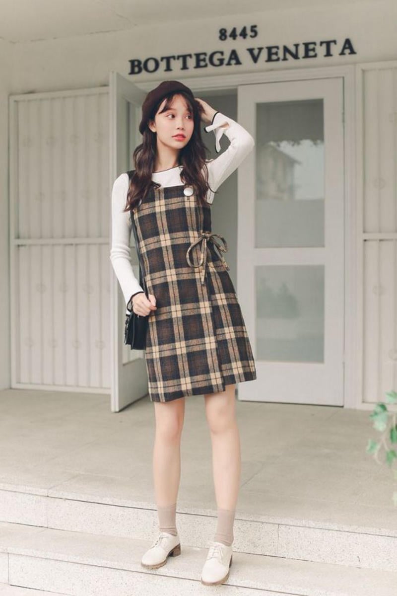 Người mẫu mặc váy kẻ caro đứng trước cửa một cửa hàng thời trang, tay cầm túi xách màu đen.