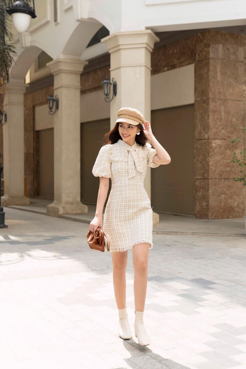 Người phụ nữ mặc váy trắng, đội mũ nâu, cầm túi, đi dạo trên phố với tòa nhà phía sau.