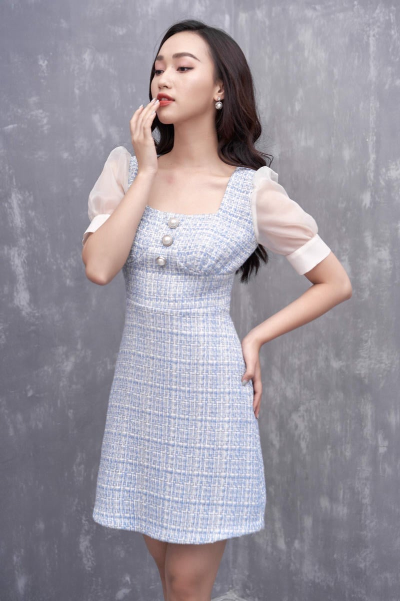 Người mặc váy xanh nhạt, tay phải đặt lên hông, tay trái gần môi, nền xám.