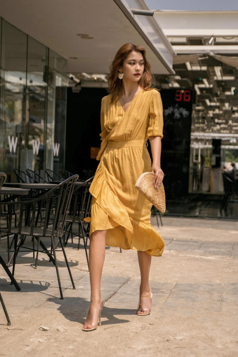 Người phụ nữ mặc váy vàng, cầm túi cầm tay, đi bộ trên vỉa hè ngoài trời, gần bàn ghế quán cà phê.