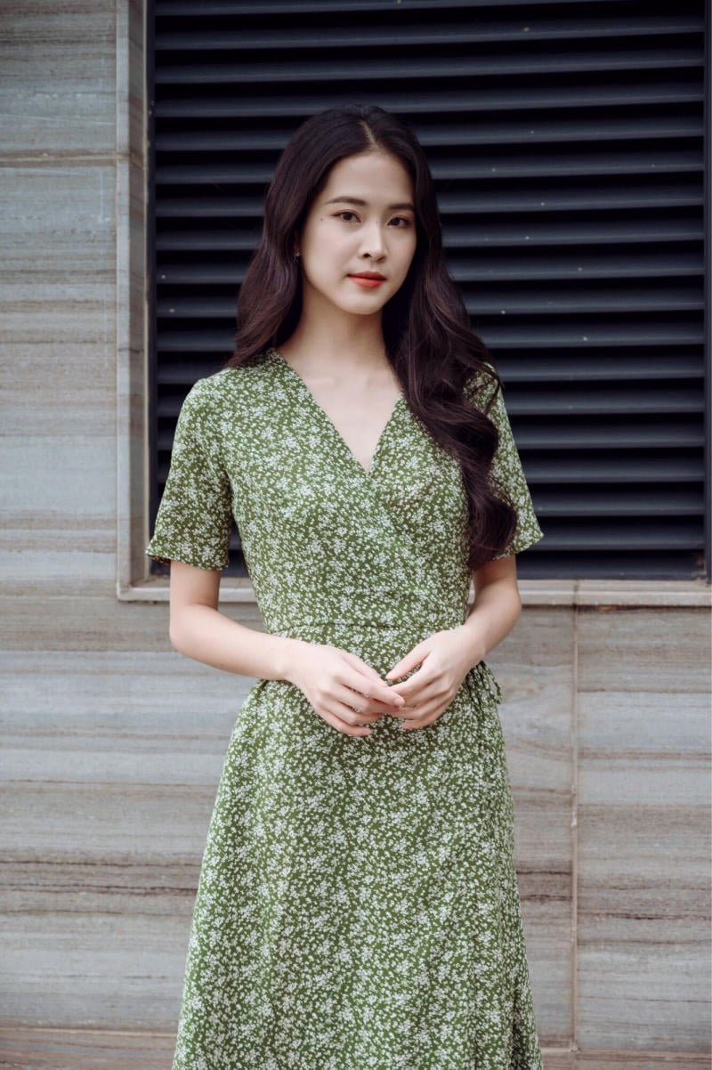 Người phụ nữ mặc váy xanh họa tiết hoa đứng trước bức tường với cửa sổ chớp đen.