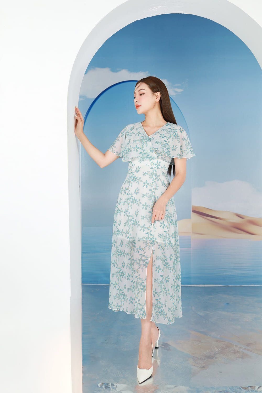 Người phụ nữ trong váy hoa xanh đứng trong khung cảnh nền trời xanh, tay chạm vào cổng vòm trắng.