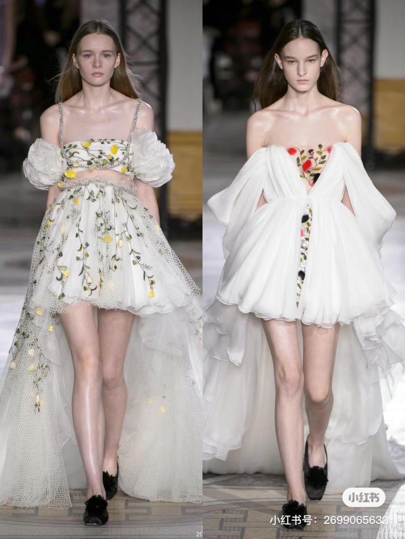 Hai người mẫu trình diễn váy trắng thiết kế cầu kỳ, trang trí hoa, trên sàn catwalk trong buổi trình diễn thời trang.