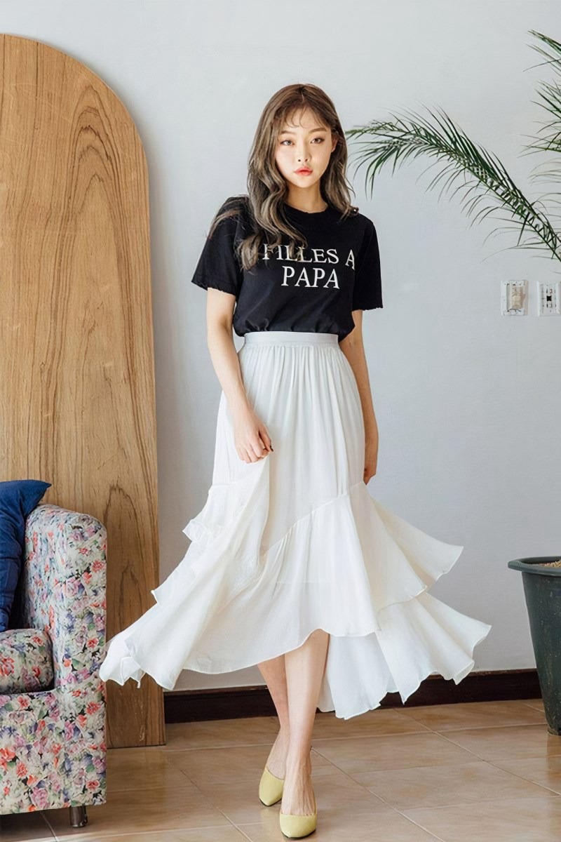 Người phụ nữ mặc áo thun đen và váy trắng dài, đứng trong phòng với cây cảnh và ghế sofa hoa.